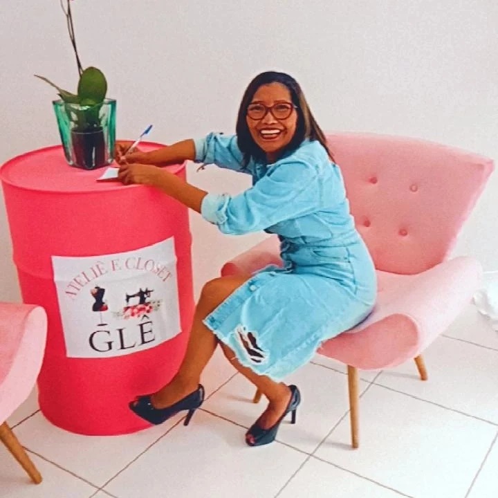 Glê sorrindo, sentada em uma poltrona rosa ao lado do tonel com a logo do ateliê